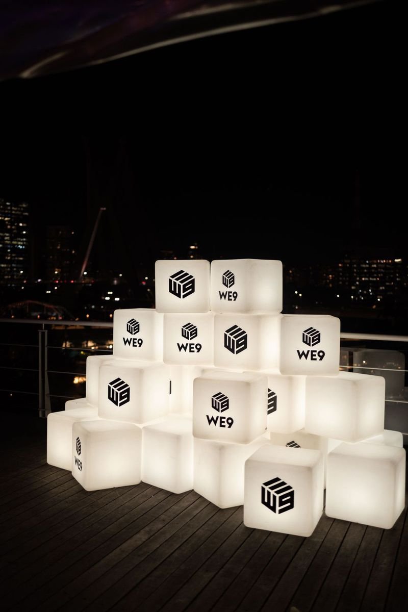 Cubos LED brancos acesos, com o logotipo WE9 em adesivo, empilhados num deck de madeira à noite, com a cidade iluminada ao fundo
