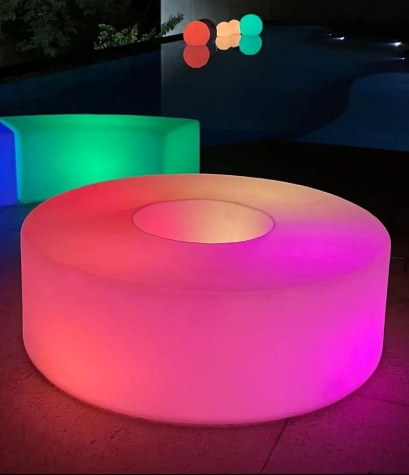 Banco LED redondo com abertura no centro, aceso em degradê de coral a rosa, à beira de uma piscina à noite
