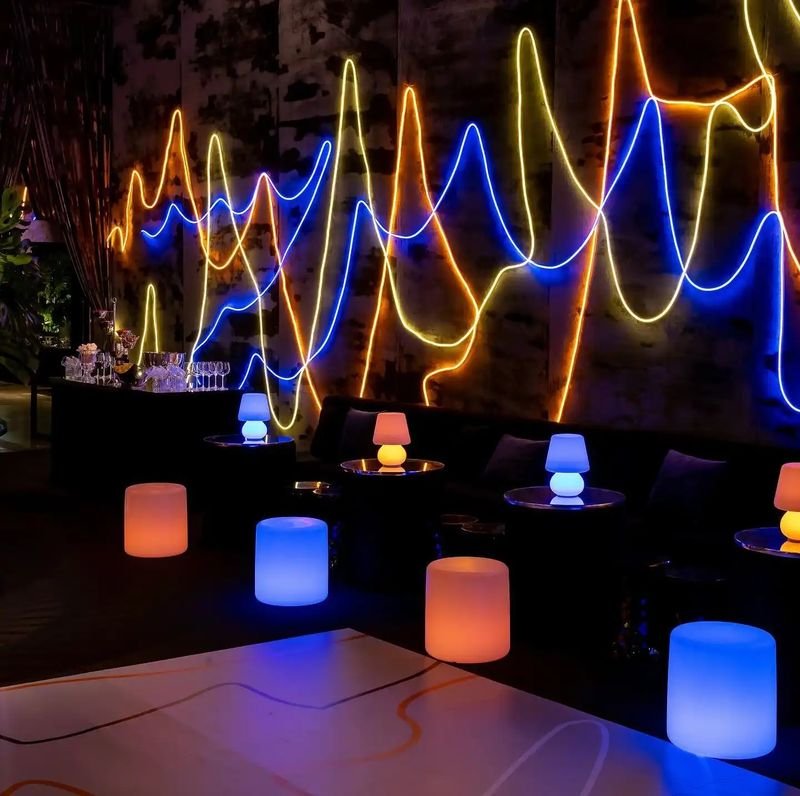 Lounge noturno com puffs LED cilíndricos acesos em laranja e azul diante de uma parede com fios de neon