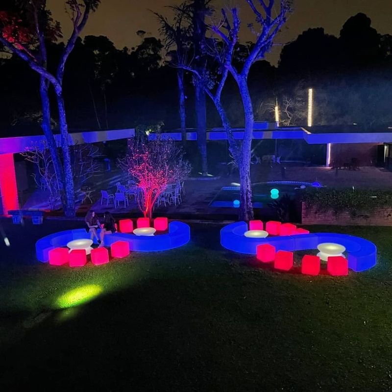 Jardim à noite com puffs LED curvos acesos em azul formando lounges, com cubos vermelhos ao redor