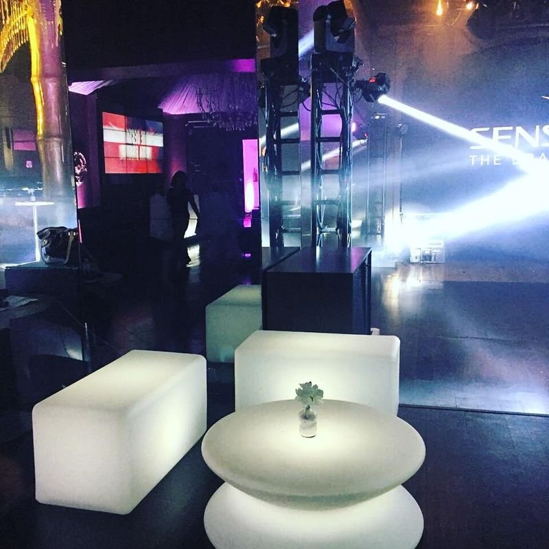 Puff LED retangular duplo aceso em branco ao lado de uma mesa iluminada, com luzes de palco ao fundo