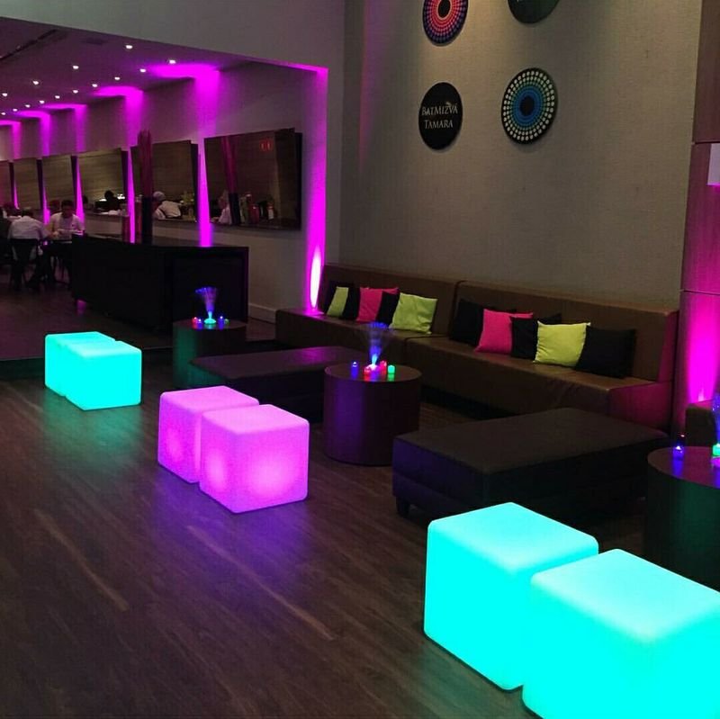 Lounge com sofás escuros e puffs LED quadrados acesos em ciano e rosa