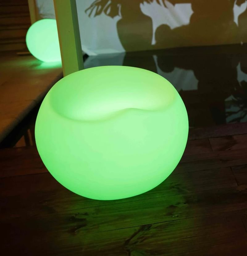 Puff LED redondo em forma de esfera, com o assento escavado no topo, aceso em verde sobre um deck de madeira, com outra esfera acesa ao fundo